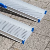 Nahaufnahme des unteren Auflagebereichs von 2 Schienen einer Aluminium-Rollstuhlrampe mit perforierter Oberfläche auf einem gefliesten Außenpflaster.