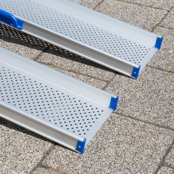 Nahaufnahme des unteren Auflagebereichs von 2 Schienen einer Aluminium-Rollstuhlrampe mit perforierter Oberfläche auf einem gefliesten Außenpflaster.