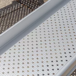 Nahaufnahme der Fahrfläche einer Rollstuhlrampenschiene mit gelochter und längsgeriffelter Oberfläche mit Unterbrechung der Riffel. Die Rampe ist aus Aluminium.