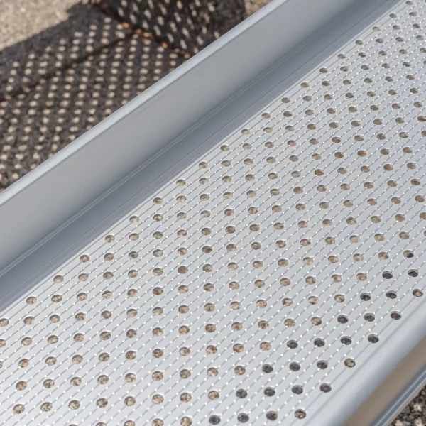 Nahaufnahme der Fahrfläche einer Rollstuhlrampenschiene mit gelochter und längsgeriffelter Oberfläche mit Unterbrechung der Riffel. Die Rampe ist aus Aluminium.