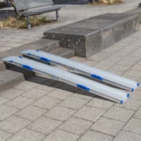 Zwei tragbare Rollstuhlrampen- Schienen liegen über einer Treppe aus 2 Stufen auf einem gepflasterten Gehweg im Freien.