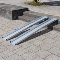 Zwei tragbare Rollstuhlrampen Schienen aus Aluminium sind über eine Außentreppe aus 2 Stufen auf einen gepflasterten Platz gelegt.