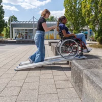 Eine Frau schiebt eine andere Frau im Rollstuhl über eine tragbare Rollstuhlrampe auf eine erhöhte Plattform im Freien.