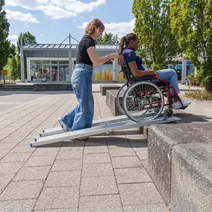 Eine Frau schiebt eine andere Frau im Rollstuhl über eine tragbare Rollstuhlrampe auf eine erhöhte Plattform im Freien.