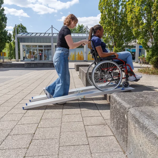 Eine Frau schiebt eine andere Frau im Rollstuhl über eine tragbare Rollstuhlrampe auf eine erhöhte Plattform im Freien.