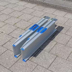 Zwei gefaltete Aluminium-Rollstuhlrampen mit blauen Griffen stehen nebeneinander auf einer gepflasterte Fläche in der Nähe eines Bordsteins.