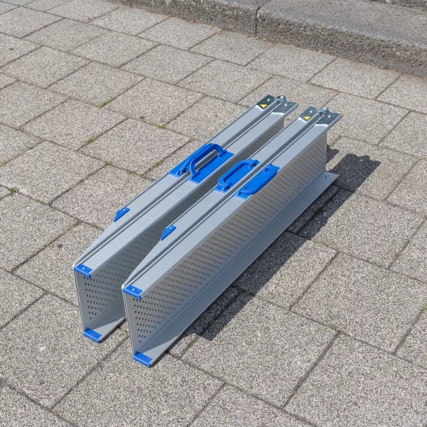 Zwei gefaltete Aluminium-Rollstuhlrampen mit blauen Griffen stehen nebeneinander auf einer gepflasterte Fläche in der Nähe eines Bordsteins.