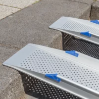 Nahaufnahme von zwei oberen Auflagewinkeln der Schienen einer Rollstuhlrampen aus Aluminium mit blauen Griffen liegen an einem sonnigen Tag im Freien auf einer Stufenkante.