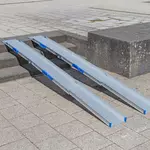 Zwei tragbare Rollstuhlrampen Schienen liegen auf einer Außentreppe und ermöglichen den Zugang zwischen verschiedenen Ebenen einer gepflasterten Fläche.