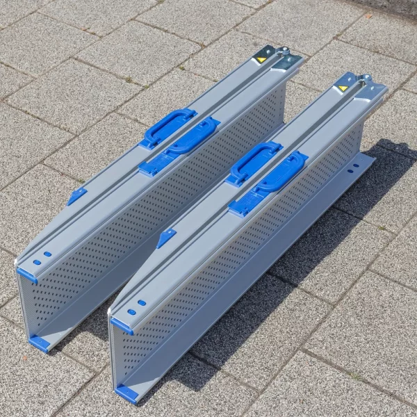 Zwei zusammen geklappte und zusammen geschobene Rollstuhlrampen Schienen aus Aluminium liegen nebeneinander auf einer gepflasterten Fläche.