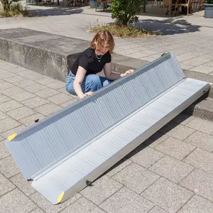 Eine Person klappt eine tragbare Aluminium-Rollstuhlrampe auf einem gepflasterten Außengelände und einer Treppe mit 2 Stufen aus.