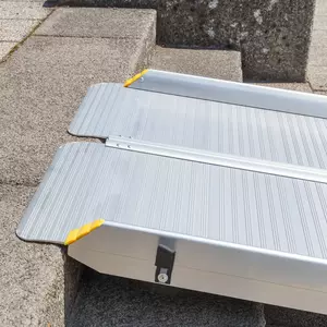 Nahaufnahme des oberen Auflagewinkels einer tragbaren Rollstuhlrampe aufliegend auf der Kante einer Betonstufe mit Darstellung der rutschsicher geriffelten Fahrfläche und des Klappscharniers.