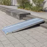 Eine tragbare Rollstuhlrampe aus Aluminium überbrückt eine kurze Außentreppe von 2 Stufen auf einem gepflasterten Gehweg.