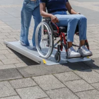 Zwei Personen benutzen eine tragbare Rollstuhlrampe; eine sitzt in einem Rollstuhl