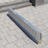 Eine zusammengefaltete tragbare Rollstuhlrampe liegt auf einer gepflasterten Fläche vor einigen Betonstufen.