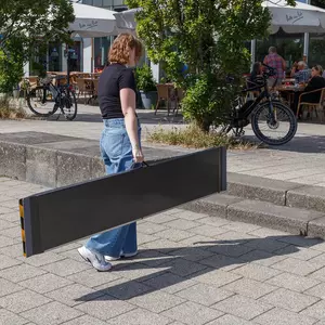 Eine Person in Jeans und schwarzem Top trägt eine schwarze Rollstuhlrampe aus Carbon auf einem Gehweg in der Nähe von Sitzgelegenheiten im Freien und Fahrrädern.