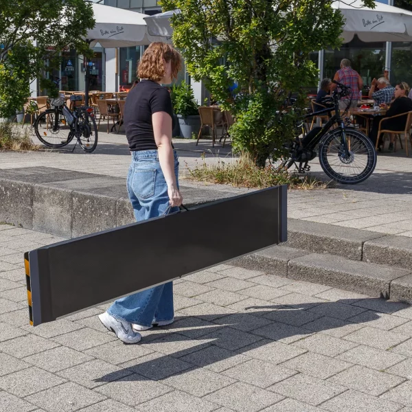 Eine Person in Jeans und schwarzem Top trägt eine schwarze Rollstuhlrampe aus Carbon auf einem Gehweg in der Nähe von Sitzgelegenheiten im Freien und Fahrrädern.