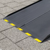 Eine Nahaufnahme des unteren Endes einer schwarzen Rollstuhlrampe aus Carbon mit gelben Warnmarkierungen am Rand auf einer gepflasterten Fläche liegend.