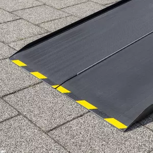 Eine Nahaufnahme des unteren Endes einer schwarzen Rollstuhlrampe aus Carbon mit gelben Warnmarkierungen am Rand auf einer gepflasterten Fläche liegend.