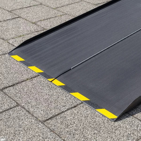 Eine Nahaufnahme des unteren Endes einer schwarzen Rollstuhlrampe aus Carbon mit gelben Warnmarkierungen am Rand auf einer gepflasterten Fläche liegend.