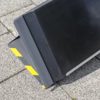 Nahaufnahme der Antirutsch- Beschichtung auf der Unterseite einer schwarzen Rollstuhlrampe aus Carbon mit einer strukturierten Kante und gelb-schwarzen Warnstreifen auf der Oberseite sowie dem Klappenscharnier auf der Unterseite.