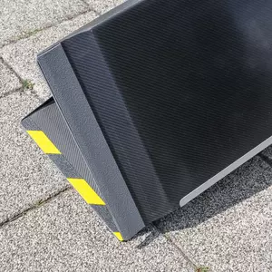 Nahaufnahme der Antirutsch- Beschichtung auf der Unterseite einer schwarzen Rollstuhlrampe aus Carbon mit einer strukturierten Kante und gelb-schwarzen Warnstreifen auf der Oberseite sowie dem Klappenscharnier auf der Unterseite.