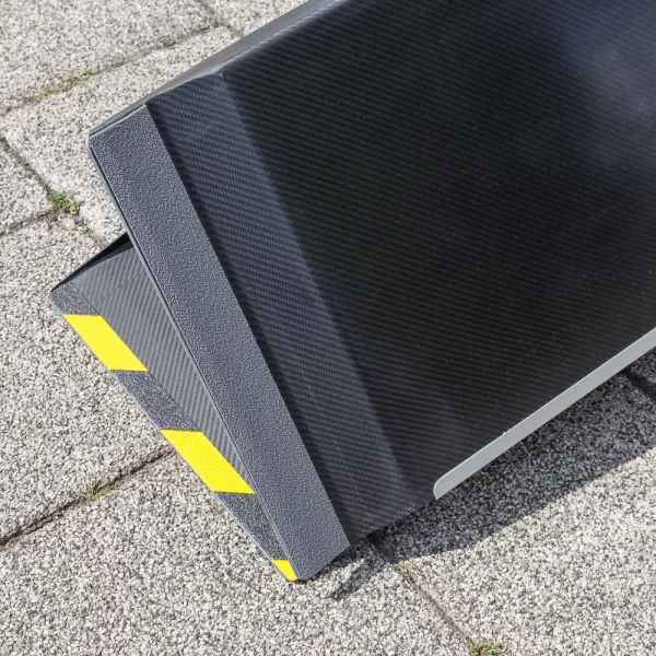 Nahaufnahme der Antirutsch- Beschichtung auf der Unterseite einer schwarzen Rollstuhlrampe aus Carbon mit einer strukturierten Kante und gelb-schwarzen Warnstreifen auf der Oberseite sowie dem Klappenscharnier auf der Unterseite.