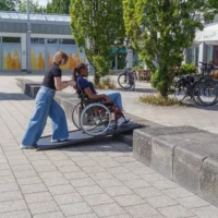 Eine Frau schiebt eine andere Frau im Rollstuhl auf einer tragbaren schwarzen Rollstuhlrampe über 2 Stufen in einem Außenbereich mit Bäumen und Fahrrädern.