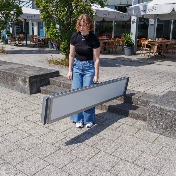 Eine Person in Jeans und schwarzem Top trägt ein faltbare Rollstuhlrampe aus Aluminium auf einem gepflasterten Außengelände in der Nähe eines Cafés.