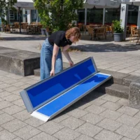 Eine Person klappt eine tragbare blaue Rollstuhlrampe / Kofferrampe aus Aluminium auf einer Außentreppe neben einem Restaurant mit Tischen und Stühlen aus.