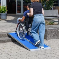 Eine Person schiebt eine andere Person im Rollstuhl auf eine tragbare blaue Rollstuhlrampe über eine Treppe im Außenbereich.