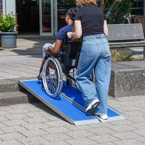 Eine Person schiebt eine andere Person im Rollstuhl auf eine tragbare blaue Rollstuhlrampe über eine Treppe im Außenbereich.