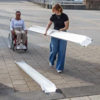 Zwei Frauen stellen eine tragbare Rollstuhlrampe auf einem gepflasterten Außengelände auf; eine Frau sitzt im Rollstuhl