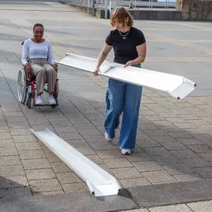 Zwei Frauen stellen eine tragbare Rollstuhlrampe auf einem gepflasterten Außengelände auf; eine Frau sitzt im Rollstuhl