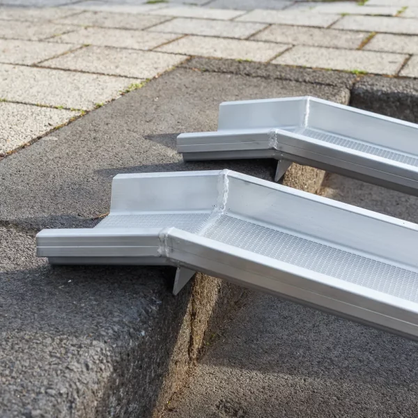 Detailaufnahme der Auflageflächen von zwei Schienen einer Rollstuhlrampe aus Aluminium mit geriffelter Fahrfläche auf der obersten Stufe einer Treppe im Freien.