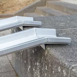 Seitliche Detailaufnahme der Auflageflächen von zwei Schienen einer Rollstuhlrampe aus Aluminium auf der obersten Stufe einer Treppe im Freien.