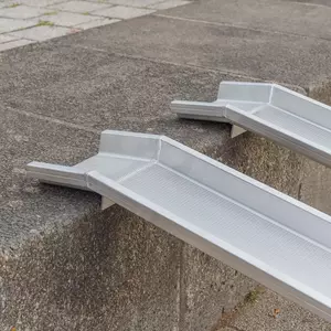 Seitliche Detailaufnahme der Auflageflächen von zwei Schienen einer Rollstuhlrampe aus Aluminium auf der obersten Stufe einer Treppe im Freien.