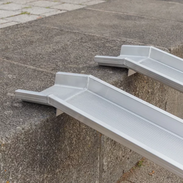 Seitliche Detailaufnahme der Auflageflächen von zwei Schienen einer Rollstuhlrampe aus Aluminium auf der obersten Stufe einer Treppe im Freien.