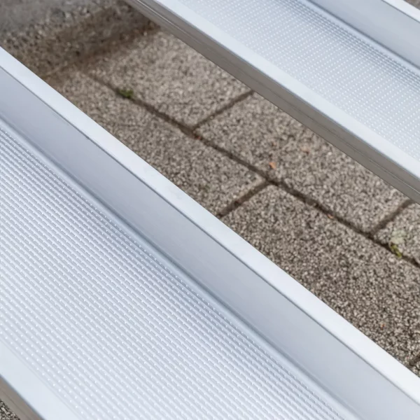 Nahaufnahme von zwei Schienen einer Aluminium-Rollstuhlrampe mit geriffelter Fahrfläche über einer Treppe im Außenbereich.