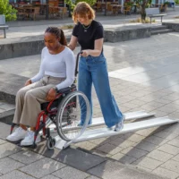 Eine Frau im roten Rollstuhl und ihre weibliche Hilfsperson am oberen Ende auf einer Schienenrampe aus Aluminium