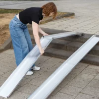 Eine Person in schwarzem Shirt und blauer Jeans platziert eine halb ausgeklappte Schiene einer Rollstuhlrampe aus Aluminium über einer dreistufigen Betontreppe im Außenbereich