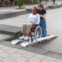 Eine Frau im Rollstuhl wird von einer anderen Frau im Freien über eine Schienenrampe geschoben.