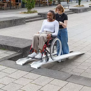 Eine Frau im Rollstuhl wird von einer anderen Frau im Freien über eine Schienenrampe geschoben.