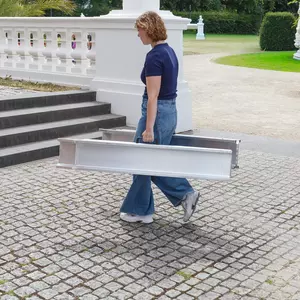 Eine Person in Jeans und blauem Hemd zwei Schienen einer zusammengeklappten mobilen Rollstuhlrampe aus Aluminium während sie draußen auf einem gepflasterten Weg geht.