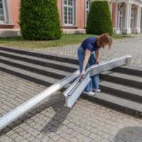 Eine Person klappt eine tragbare Rollstuhlrampe auf einer Steintreppe in der Nähe eines Gebäudes mit großen Fenstern und beschnittenen Büschen aus. Eine Schiene der Rampe liegt bereits aufgebaut über den Treppenstufen.