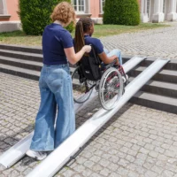 Eine Frau schiebt eine Frau im Rollstuhl über die zwei parallel platzierten Schienen einer Schienenrampe aus Aluminium von unten nach oben über eine Treppe mit vier Stufen im Außenbereich