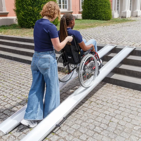 Eine Frau schiebt eine Frau im Rollstuhl über die zwei parallel platzierten Schienen einer Schienenrampe aus Aluminium von unten nach oben über eine Treppe mit vier Stufen im Außenbereich