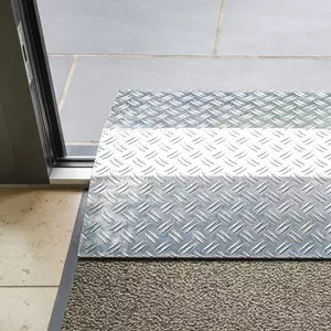 Eine Rollstuhlrampe aus Aluminium mit geriffelter Oberfläche führt von einer Fußbodenmatte zu einem gefliesten Außenbereich.