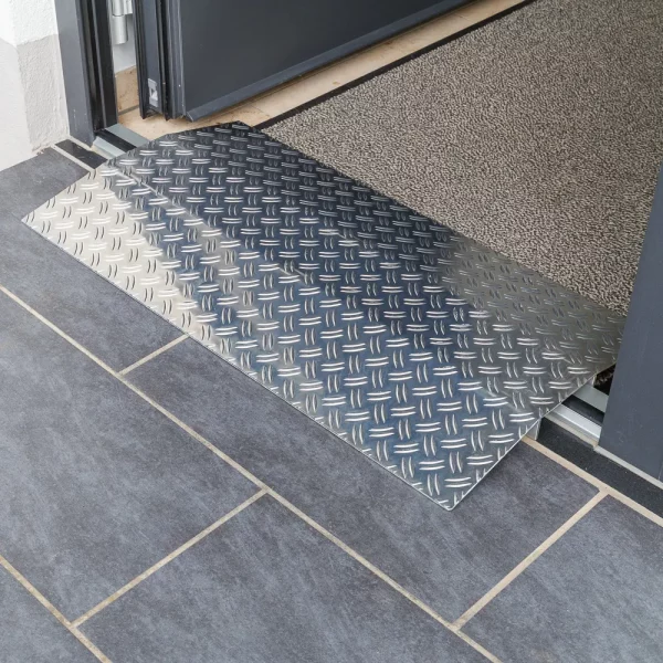 25 Rollstuhlrampe Schwellenbrücke diagonal rechts Eine Schwellenrampe aus Aluminium überbrückt eine Türkante und einem gefliesten Boden und einem mit Fußmstte ausgelegten Raum innerhalb einer Türöffnung.