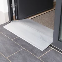 Eine Schwellenbrücke aus Aluminium liegt an der Schwelle einer Türöffnung über dunklen Bodenfliesen neben einer Fußmatte.